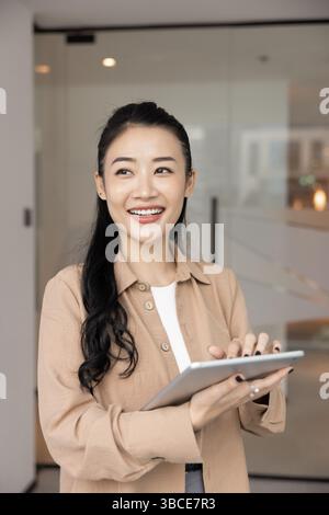 Smiling Chinese businesswoman holding tablet device standing in modern workplace Stock Photo