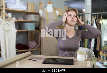 Tired female fashion designer sitting at table in sewing studio Stock Photo