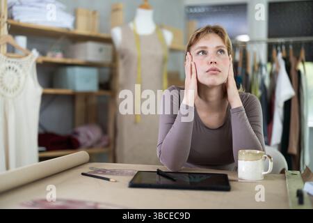 Tired female fashion designer sitting at table in sewing studio Stock Photo