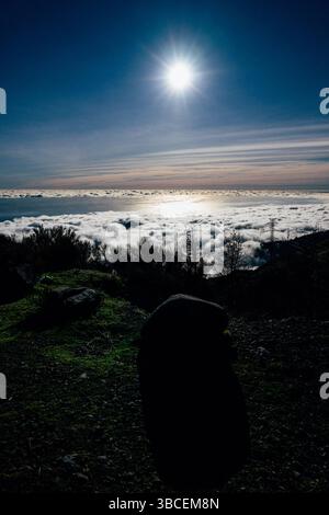 Bright sun shines over a sea of clouds from a mountain viewpoint at dusk Stock Photo