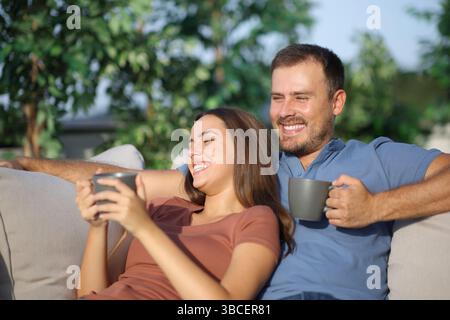 Happy couple sitting in a sofa in the garden drinking coffee at home Stock Photo