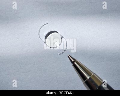A close-up view of a transparent lens placed on textured paper, with a silver pen tip in the foreground, creating a contrast between the lens and the Stock Photo