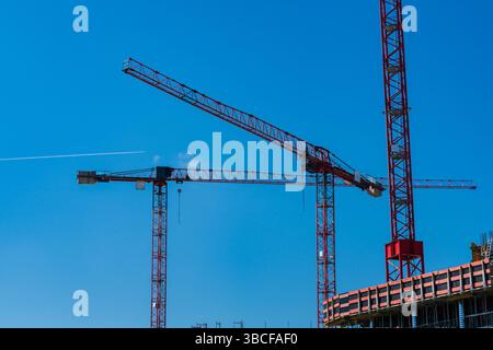 Large red construction cranes stand tall against a clear blue sky, actively engaged in lifting materials for a new building project. A plane streaks p Stock Photo
