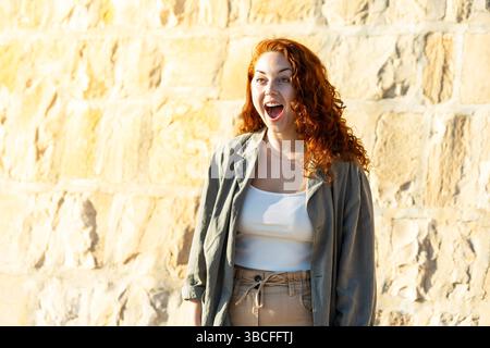 Excited young woman with curly red hair showing a surprised facial expression, mouth wide open, standing against a bright sunlit stone wall background Stock Photo
