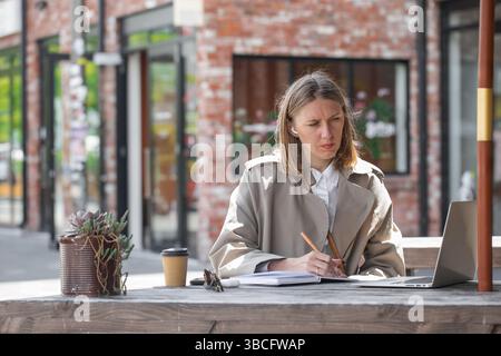 Woman taking notes in notebook during online meeting in open air cafe. Businesswoman  Stock Photo
