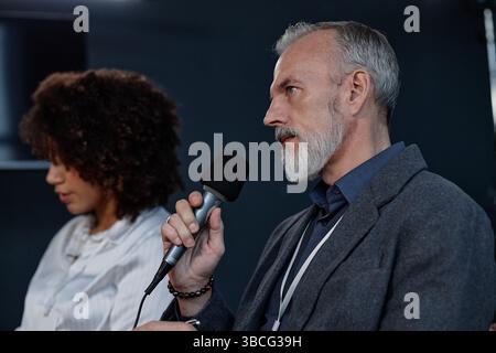 Side view of mature male investor holding microphone asking executives questions about financial performance while sitting in audience space during AG Stock Photo