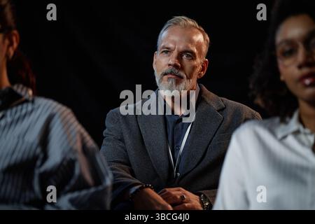 Medium shot of focused senior male conference participant dressed in suit listening to speakers presentation, while attending business seminar in audi Stock Photo