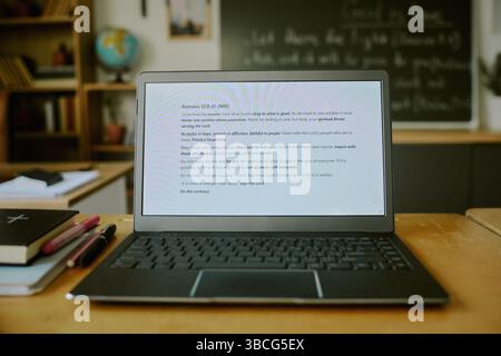 Laptop displaying Bible passage, open on classroom desk with books and stationery nearby, and blackboard in background. Scene depicting serene study e Stock Photo