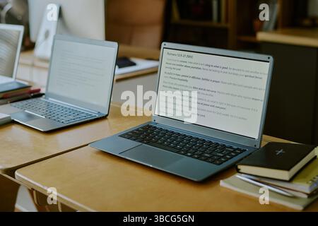 Modern workspace with pair of laptops displaying on screen text, situated on wooden desks. Background includes books, papers, and blurred office furni Stock Photo