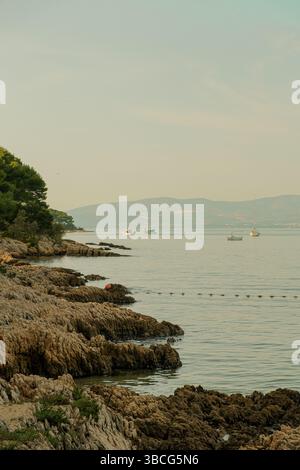Scenic coastal view with sailboats on calm sea, framed by pine tree branches, with mountains and a seaside town in the background. Stock Photo