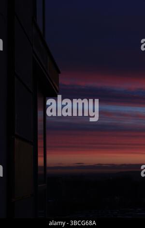 A dark apartment building facade reflects the last light of a vibrant sunset, with streaks of pink, purple, and blue clouds stretching across the sky Stock Photo