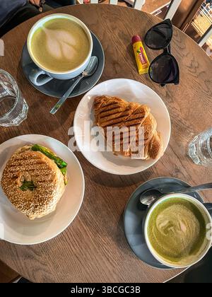 Top-down view of a wooden table in a cozy café setting, with two matcha lattes, a sesame bagel sandwich with lettuce, and a flaky croissant. Stock Photo