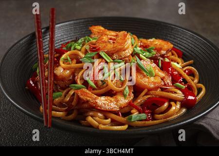 Udon stir-fry shrimp noodles with red bell pepper and green onion, served in a black bowl, with chopsticks, dark background Stock Photo
