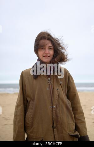 Woman in a brown coat stands on a windy beach with overcast skies in the background. Ostend, Belgium Stock Photo
