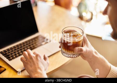 A handsome man relaxes at a table with a drink while focusing on his laptop in a lively area. Stock Photo