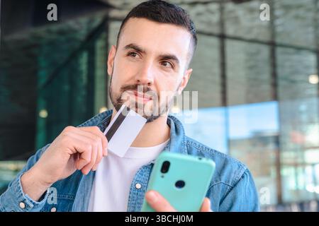 Thoughtful man holding credit card and smartphone in modern urban environment Stock Photo