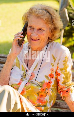 Elderly woman with glasses hanging from a string around her neck is sitting on a wooden bench outside in a garden, smiling while talking on her smartp Stock Photo