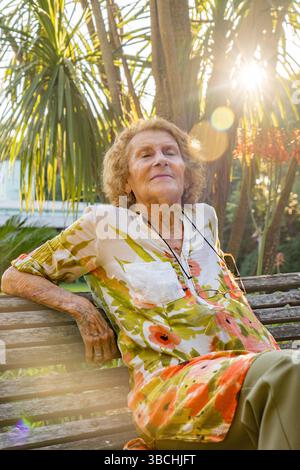 Elderly woman with closed eyes enjoying sunlight while sitting on wooden bench in lush botanical garden, embracing tranquility and nature's beauty Stock Photo