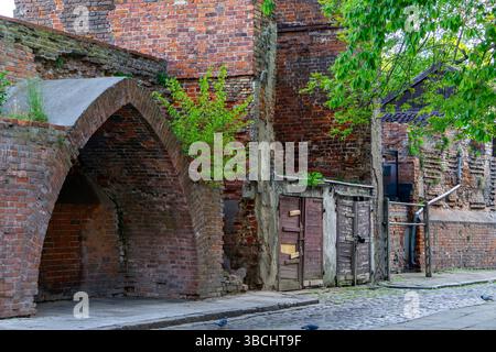 Historic section of urban brick architecture featuring an arched passage, old wooden doors, and a partially ruined facade. Overgrown greenery adds cha Stock Photo