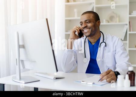 Smiling Black Male Doctor Talking On Cellphone And Using Computer At Workplace Stock Photo