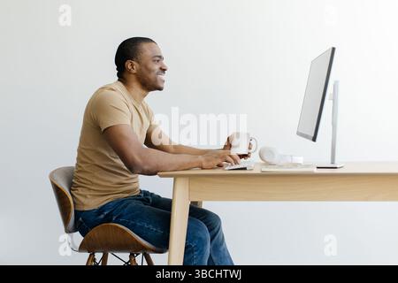 Smiling African American man working on desktop computer and drinking coffee Stock Photo