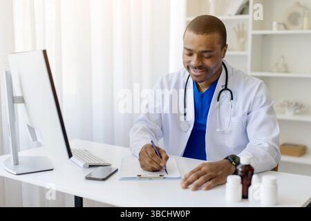 Happy black doctor man sitting at desk in clinic and taking notes Stock Photo