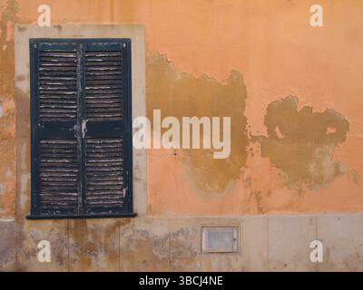 An old window with green painted closed wooden shutters on an orange Mediterranean ocher colored orange cracked peeling flaking wall Stock Photo