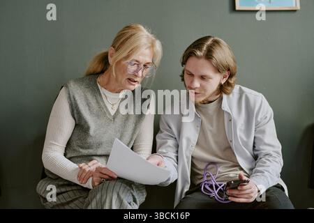 Elderly woman wearing glasses holding papers and interacting with young man holding gadget, engaged in conversation, sitting on a couch against green Stock Photo