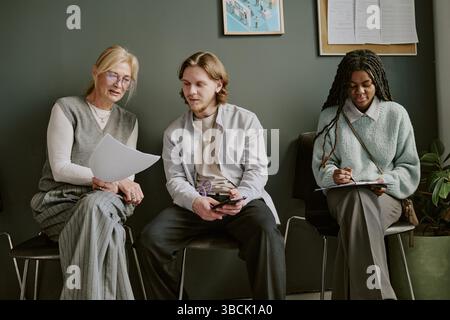 Group of colleagues brainstorming in an office setting, sitting on chairs and engaging in discussion while referencing documents, with a diverse team Stock Photo