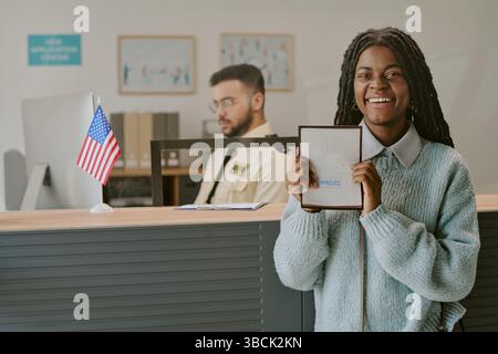 Smiling African American woman holding documentation while standing in front of reception desk and American flag, with man working in background Stock Photo
