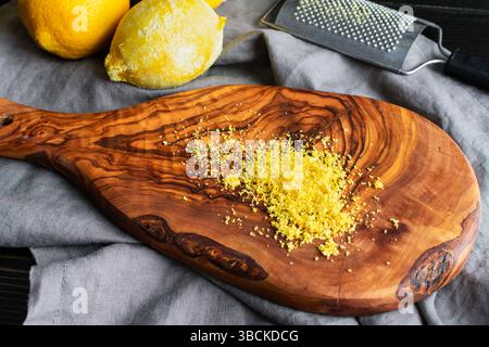 Lemon Zest on an Olive Wood Cutting Board: Lemon zest with zester grater and fresh lemons in the background Stock Photo