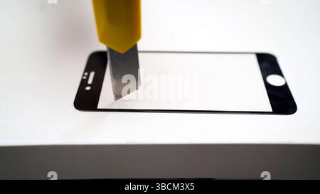 Phone repair technician is using a sharp utility knife to remove a cracked tempered glass screen protector from a smartphone Stock Photo