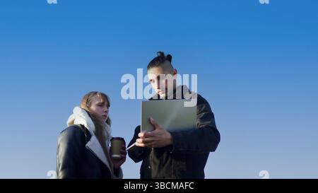 Two young professionals collaborating outdoors, using laptop and drinking coffee under blue sky Stock Photo