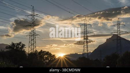 sunset in a mountain landscape with electrical power lines and lattice crosses in the foreground and a setting sun disappearing behind silhouette of m Stock Photo