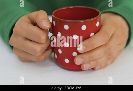 delicate female hands of a woman in a bright green sweater holding an empty red coffee cup with white polka dots on a white tablecloth Stock Photo