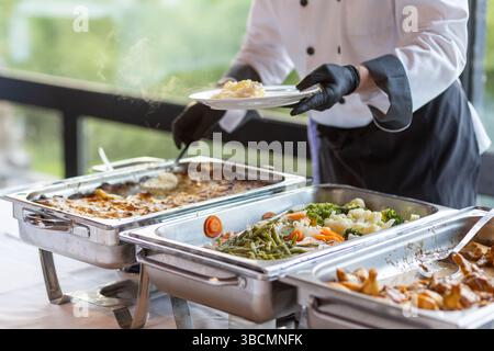 A chef in a black and white uniform carefully serves a plate of food from a buffet at an outdoor event in the afternoon. Various dishes are displayed, Stock Photo