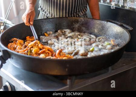Fresh shrimp and other seafood sizzle in a large pan over a flame at a vibrant outdoor market. The aroma fills the air as a cook skillfully stirs the Stock Photo