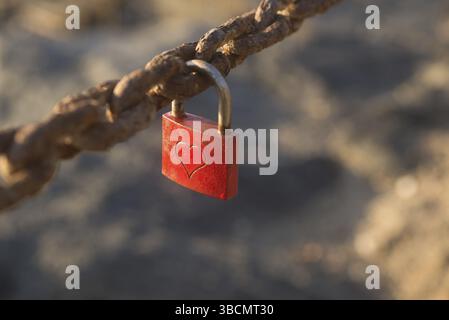 A small lock, painted red, with a heart engraved on it, is locked on to a rusty chain Stock Photo