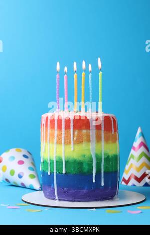 Delicious rainbow birthday cake with burning candles, party hats and confetti on light blue background Stock Photo