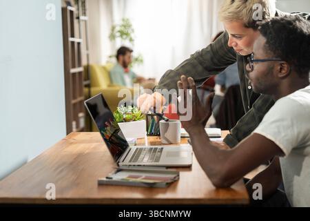 In a bright home office setting, a Black man and a female colleague lean over a laptop, pointing at the screen and collaborating with smiles during a Stock Photo
