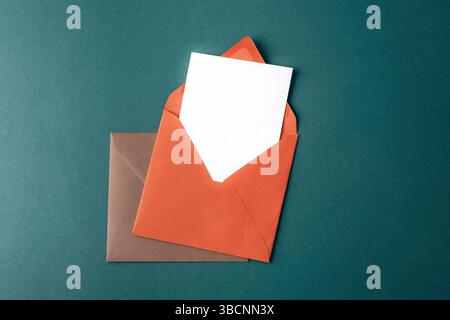 Orange and brown envelopes with blank white card on dark green background. Top view, flat lay, mockup. Stock Photo
