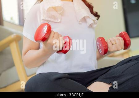 Cropped shot of sporty mature woman with red dumbbells on light room. Mature woman training with dumbbells at home. Selective focus. Stock Photo