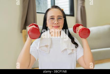 Portrait of cheerful aged Asian woman in fitness wear exercising with red dumbbells in livingroom at home. Elderly woman prefers healthy lifestyle con Stock Photo