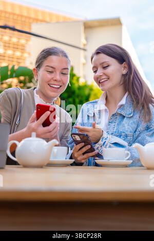 Two caucasian women using smartphones while enjoying tea at outdoor cafe table Stock Photo