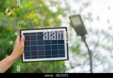 Cropped shot of man hand holding a solar panel with blurred background of a metal pole of streetlamp. Sustainable energy in daily life concept. Stock Photo