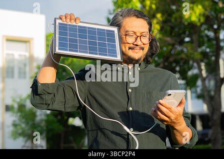 Handsome man with eyeglasses using mobile phone charging from portable solar panel at public city park. Selective focus. Stock Photo