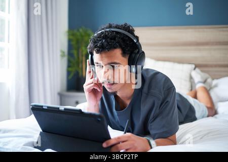 Young Man Using Tablet While Relaxing with Headphones on Bed at Home Stock Photo
