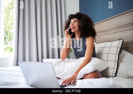 Smiling Young Woman Sitting in Bed While Talking on Phone and Using Laptop Stock Photo