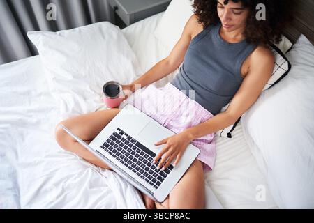 Woman Relaxing on a Bed While Using a Laptop and Drinking Beverage Stock Photo