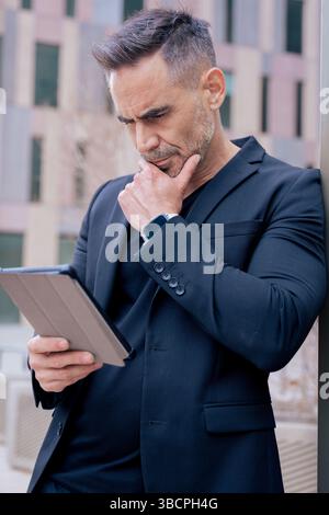 Thoughtful mature man using tablet while leaning on pole in city.Vertical Stock Photo